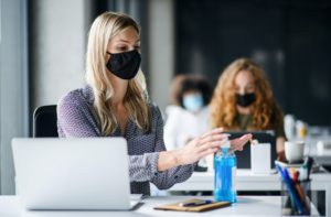 Young woman with face mask back at work in office after lockdown, disinfecting hands
